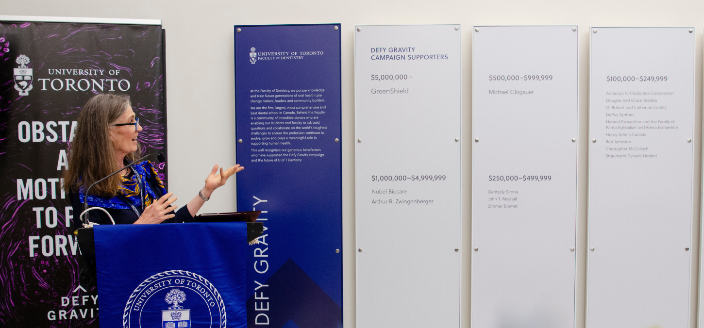 Photo of Elizabeth MacSween speaking next to the donor wall. 