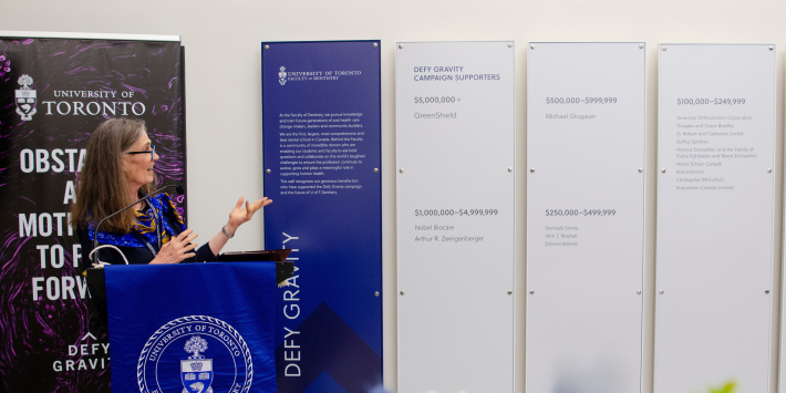 Photo of Elizabeth MacSween speaking next to the donor wall. 